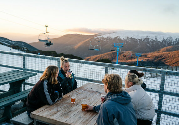 A group of five young adults sit at a wooden picnic table drinking beers at a venue perched on top of a mountain. They admire views of Falls Creek's snow-covered Mountain scape at sunset.