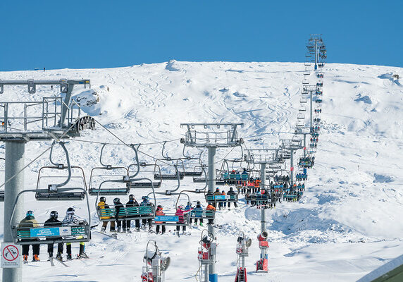 A wide shot of ruin castle chairlift spanning from the bottom of the mountain to the top. The chairlifts are full of people looking out at fresh tracks on a sunny blue-sky day.